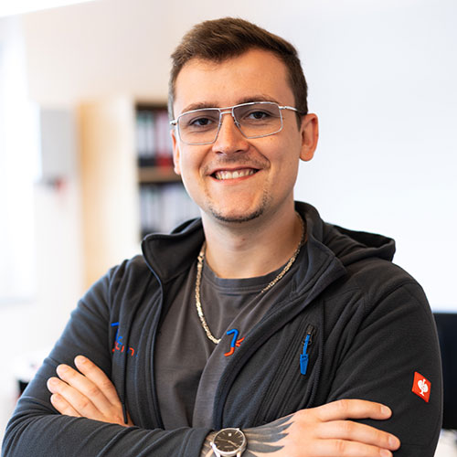 Mann mit Brille lächelt, trägt blaue Jacke mit Logos, Tattoo am Handgelenk sichtbar.