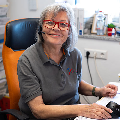 Ältere Frau mit grauem Haar und roter Brille lächelt, trägt ein graues Polohemd und ein Headset.